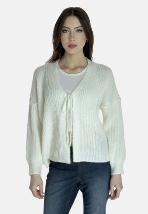 Weißer gestrickter Cardigan mit V-Ausschnitt, gebundenem Verschluss und Ballonärmeln. Die Struktur ist dick und weich und wird mit dunkelblauen Jeans kombiniert.