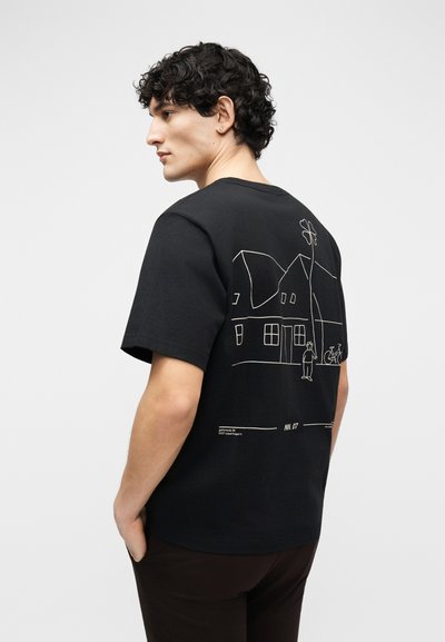 Νεαρός άνδρας με σγουρά μαλλιά φορώντας μαύρο μπλουζάκι με απεικόνιση λευκής γραμμικής τέχνης ενός σπιτιού, ενός ατόμου και ενός ποδηλάτου στην πλάτη, κοιτάζοντας προς τα αριστερά.