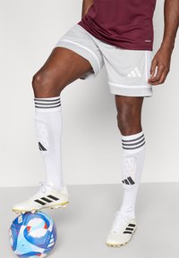 Weiße Fußballschuhe mit schwarzen Akzenten, graue Shorts mit weißen Streifen und kniehohe weiße Socken mit schwarzen Streifen und Logos.
