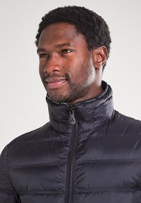 Giubbotto imbottito nero con colletto alto, caratterizzato da cuciture orizzontali, una cerniera frontale completa e un accento del colletto con logo.