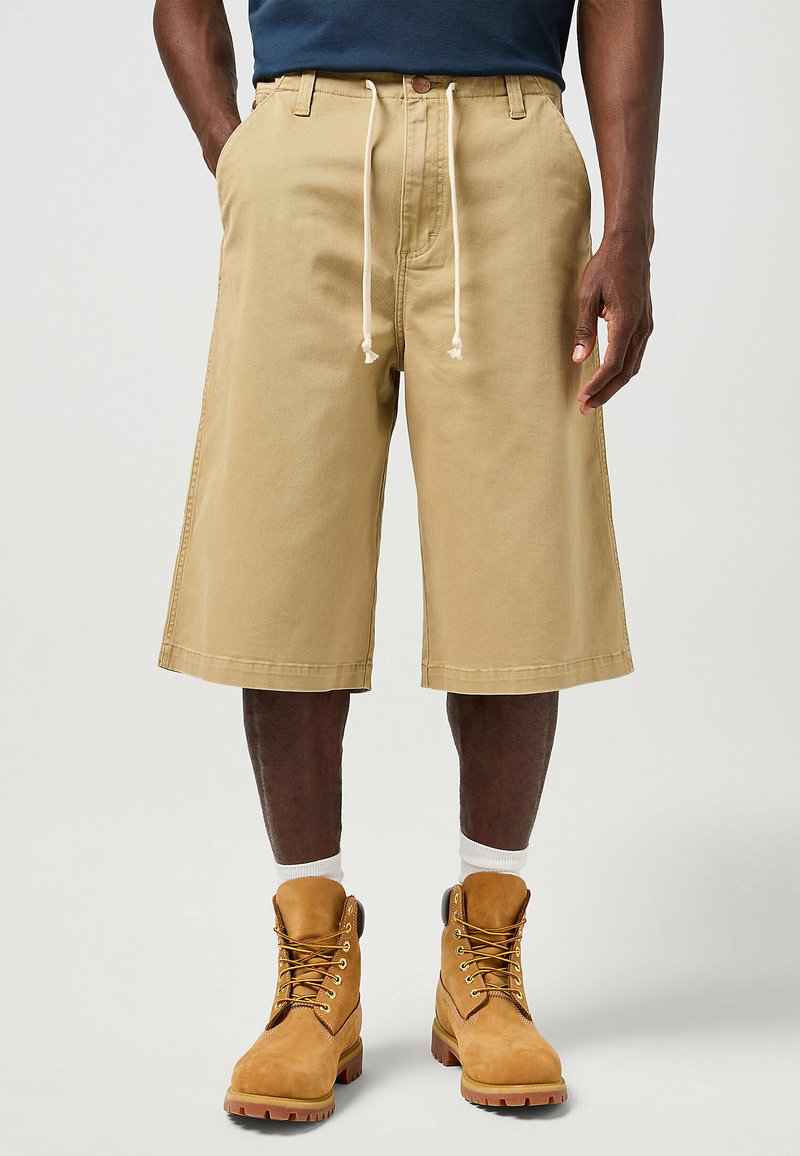 Beige shorts lavet af bomuld med en løs pasform, featuring en elastisk talje og sidelommer. Kombineret med brune arbejdssko og hvide sokker.