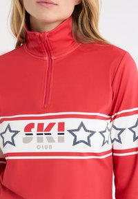 Czerwony sportowy top zapinany na zamek z białymi i granatowymi paskami oraz napisem „SKI CLUB”. Materiał o fakturze z półgolfem.