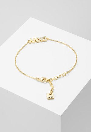 Bransoletka z łańcuszkiem w kolorze złota z zawieszką z wygrawerowanym napisem „DKNY” oraz ozdobnymi literami „DKNY” z kryształowymi akcentami na białej powierzchni ekspozycyjnej.