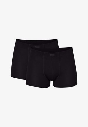 Oberkörperfreier Mann mit langem, dunklem, zurückgebundenem Haar, der schwarze Boxershorts trägt und barfuß vor einem schlichten hellen Hintergrund steht.