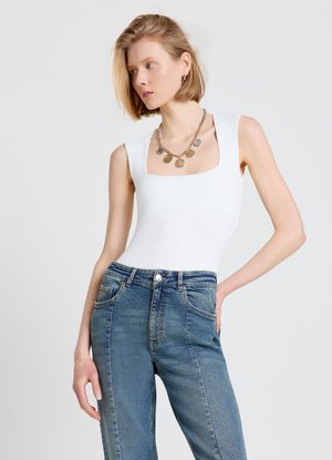 Femme aux cheveux blonds courts porte un haut blanc sans manches, un jean bleu taille haute et un collier avec une pièce en or, posant la main sur la hanche.