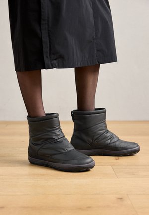 Fekete párnázott bokacsizmák, texturált gumitalppal és függőleges varrásokkal. A dizájn egy hajtott gallért tartalmaz a megnövelt melegítés érdekében.