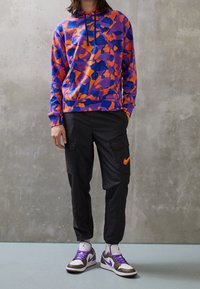 Fialová mikina s kapucí se abstraktními vzory v oranžové a modré barvě, doplněná černými cargo kalhotami s červeným logem Nike a hnědo-bílými teniskami.