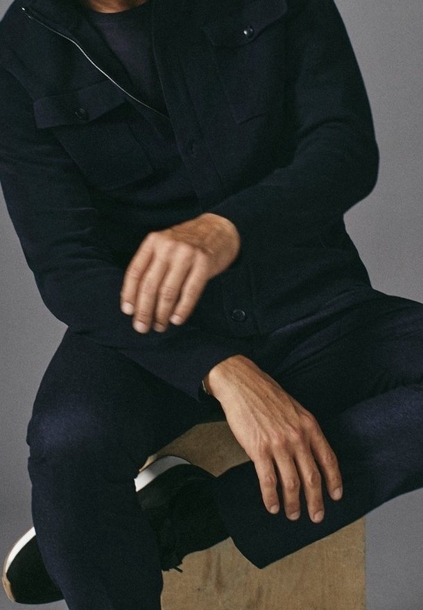 En man med svart jacka och byxor sitter på en träblock, med ena handen vilande på knät och den andra i rörelse över knät.