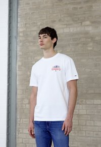 Hvid bomulds T-shirt med rød "TOMMY'S" print og blå accenter, parret med blå denim jeans, stående op ad en lys mursten væg.