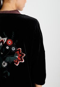 Femme portant une veste en velours noir avec de la broderie florale rouge et blanche dans le dos, tournée légèrement sur le côté.
