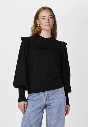 Maglione nero con dettagli a volant sulle spalle e sul petto, polsini aderenti e scollatura a coste. Abbinato a jeans in denim blu chiaro.