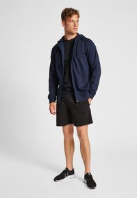Felpa con zip colore navy con cordini, abbinata a una t-shirt nera e pantaloni corti neri. Il modello indossa scarpe da ginnastica nere, in piedi su uno sfondo neutro.