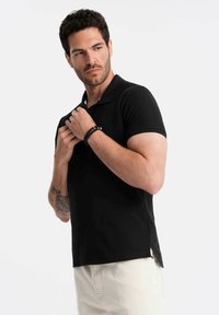 Mann in schwarzem Poloshirt und weißer Hose richtet den Kragen, trägt ein schwarzes Perlenarmband und hat ein sichtbares Tattoo am linken Unterarm.