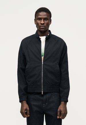 Mladý muž nosi čiernu bundu na zips cez biele tričko s potlačou, stojí na jednoliatom svetlom pozadí.