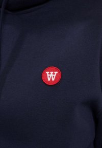 Marineblå hettegenser med strukturert stoff som har en sirkulær rød lapp, med en hvit "W"-logo. Enkel design, ingen andre synlige detaljer.