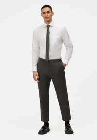 Camicia bianca con taschino, cravatta verde scuro e pantaloni grigi scuri a gamba affusolata. Scarpe nere con suola spessa. Tessuto morbido e vestibilità sartoriale.