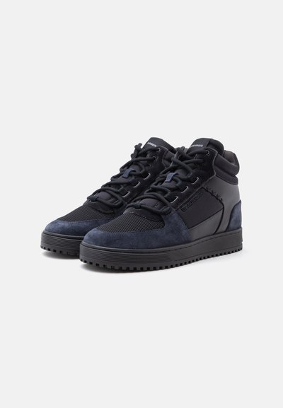Ζευγάρι μαύρα και σκούρα μπλε sneakers ψηλού τύπου με κορδόνια, υφασμένους ενισχυμένους μπροστινούς τμήματα και επενδεδυμένα περιλαίμια αστραγάλου σε απλό φωτεινό φόντο.