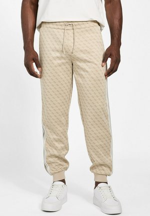 Pantaloni jogger beige con motivo, strisce laterali bianche, polsini elasticizzati e vita con coulisse, indossati con sneakers bianche da una persona in piedi.