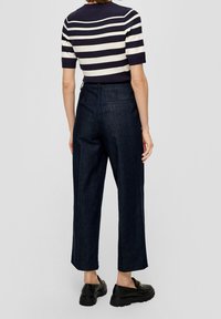 Person klädd i svarta loafers, mörka högmidjade vida byxor och en svart-vit horisontellt randig kortärmad topp, stående mot en grå bakgrund.