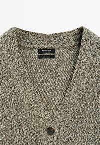 Nahaufnahme eines beigefarbenen und grauen gestrickten Baumwollpullovers mit V-Ausschnitt und einem einzelnen dunklen Knopf, der den Text des Innenlabels zeigt.