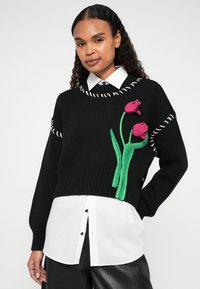 Sweater preto de malha com design oversized e costura branca, decorado com bordados de flores verdes e rosas. Usado por cima de uma camisa branca.