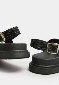 Mustat korokepohjatossut, joissa on teksturoitu pohja, säädettävä remmi kultaisen sävyn soljella ja kontrastivalkoinen ommel remmeissä.