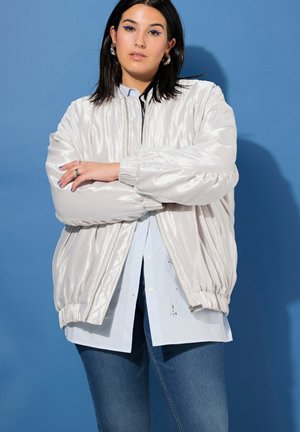 Una mujer con los brazos cruzados luce una brillante cazadora bomber blanca sobre una camisa clara y unos vaqueros, delante de un fondo de estudio azul.