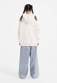 Krémová oversized mikina s kapucí, stylizovaná s volnými, lesklými stříbrno-modrými kalhotami s jemnou texturou a bočními akcenty.