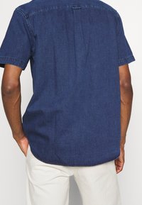 Camicia in denim scuro a maniche corte con orlo curvo e piega sul retro, caratterizzata da una finitura pulita e semplici dettagli di cucitura.