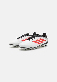 Botas de fútbol blancas con acentos rojos, cordones negros y suela negra/blanca. Presentan una parte superior sintética suave y un destacado logo de tres rayas.