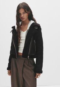 Giacca nera cropped con colletto in shearling, dettagli in cerniera argentata e finitura strutturata. Indossata sopra un hoodie grigio chiaro e pantaloni marroni.
