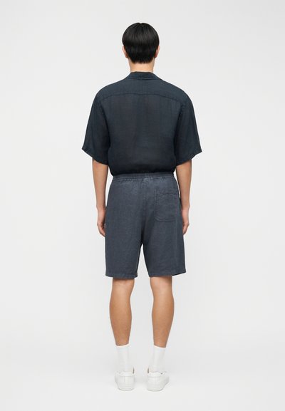 Homme portant une chemise à manches courtes foncée, un short gris avec une poche arrière, des chaussettes blanches et des baskets blanches, debout face à un fond blanc uni.