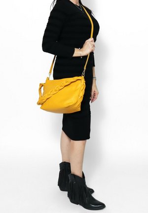 Gelbe Leder-Schultertasche mit geflochtenem Griff, lässiger Form und verstellbarem Riemen, getragen mit einem schwarzen Kleid und gefransten schwarzen Stiefeletten.