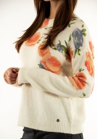 Mujer vistiendo un suéter de punto color crema con patrones florales en naranja y azul, con las manos entrelazadas y pantalones oscuros, frente a un fondo blanco.
