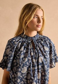 Námornícky modrá kvetinová blúzka so zhrnutým výstrihom, krátkymi volánovými rukávmi a očkovým zapínaním vpredu, zdobená bielymi botanickými vzormi.
