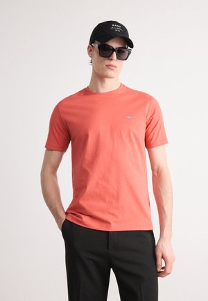 Korallenfarbiges Baumwoll-T-Shirt mit kurzen Ärmeln und Rundhalsausschnitt, mit einem kleinen Logo auf der linken Brust. Getragen mit schwarzen Hosen und Zubehör.