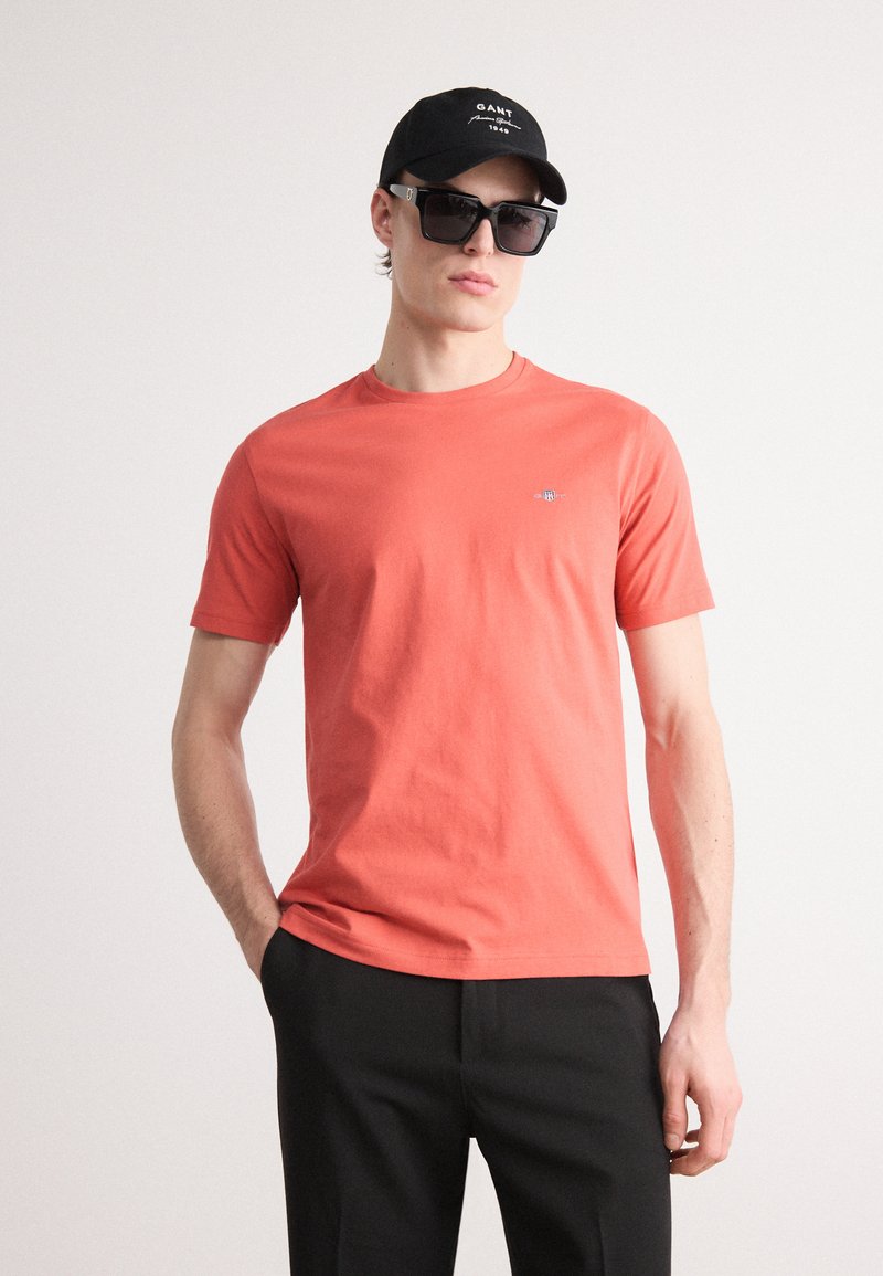Tricou din bumbac coral cu mâneci scurte și decolteu rotund, având un mic logo pe pieptul stâng. Poartă pantaloni negri și accesorii.