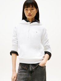 Mujer joven vistiendo una sudadera blanca sobre una camisa de mezclilla negra y pantalones de mezclilla negra, con una mano en el bolsillo, frente a un fondo liso.
