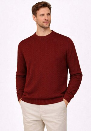 Uomo che indossa un maglione a maniche lunghe color borgogna con trama e pantaloni beige chiaro, in piedi con le mani nelle tasche, guarda verso destra.