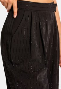 Zwarte geplooide broeken met metallic streepdetails. Gemaakt van een gladde stof, met een gestructureerde tailleband en zijzakken.
