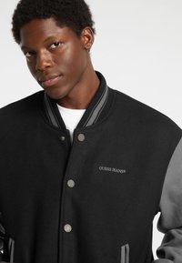 Čierna varsity bunda s šedými akcentmi, so zapínaním na patentky a vyšívaným logom "GUESS JEANS" na ľavej hrudi. Materiál z vlnenej zmesi.