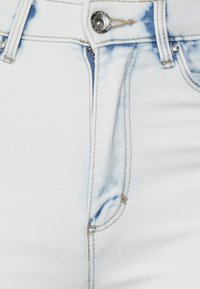 Tecido de denim branco com detalhes em azul claro desgastados. Apresenta um fecho padrão, botão de metal e costura visível ao longo das costuras.