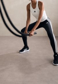 Personne en débardeur blanc et leggings Adidas noirs faisant des exercices avec des cordes de combat sur un sol gris avec un mur beige.