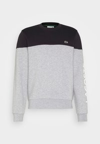 Szara bluza z czarną górną częścią, ściągaczami na mankietach oraz białym logo "LACOSTE" na rękawie. Miękki, średniej grubości bawełniany materiał.