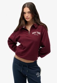Bordowy krótki sweatshirt z zamkiem w kołnierzu, z białym napisem "NEW YORK". W połączeniu z granatowymi dresami, prezentującymi casualowy styl.