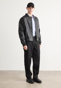 Black faux leather bomber jacket with quilted accents. Light blue shirt underneath, paired with loose black trousers and black shoes.
