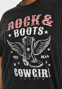Czarna koszulka z napisem "ROCK & BOOTS" w paski czerwono-białe, graficzną ilustracją kowbojskich butów z skrzydłami oraz napisem "YEE HAA COWGIRL" białymi literami.