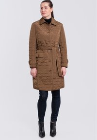 Abrigo acolchado marrón con cuello puntiagudo, cierre de botones y cinturón. Superficie texturada con formas de diamante, combinado con jeans oscuros y botas de tobillo.