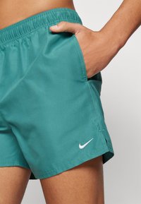 Shorts de bain teal avec une taille élastique, poches latérales et un logo Nike blanc sur l'ourlet ; tissu lisse avec un design léger.
