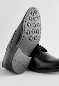 Chaussure en cuir noir avec une finition lisse, dotée d'une semelle en caoutchouc grise avec des points en caoutchouc ronds pour l'accroche. Design à lacets.